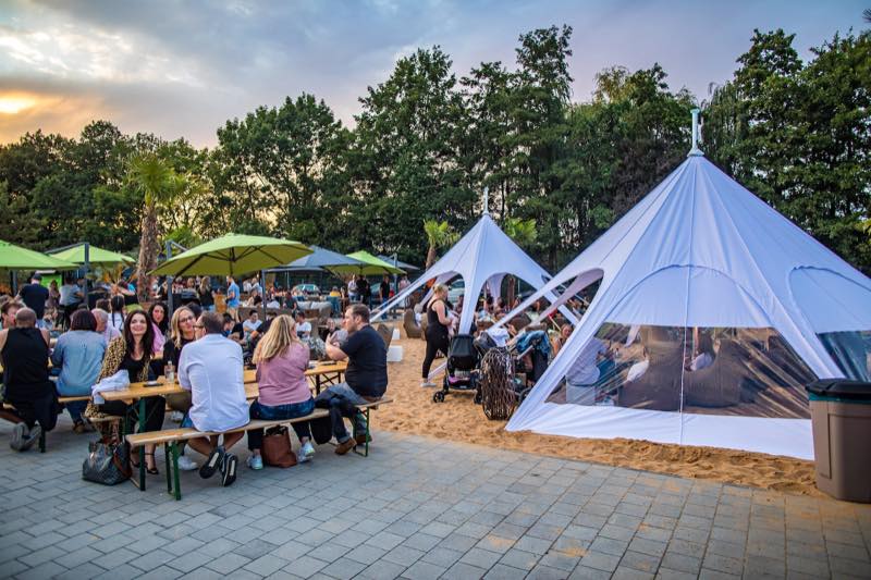 Gäste an Biertischen im Sand, dahinter Sternzelt und Sonnenuntergang